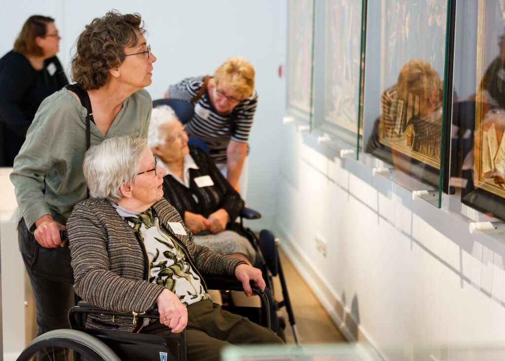twee oudere dames in rolstoel met elk een vrouwelijke begeleider bekijken aandachtig een tentoonstelling achter glas aan de wand in Rijksmuseum Boerhaave